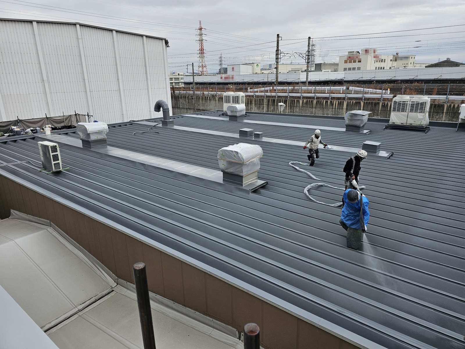 一般部の吹付施工中。軒先から奥の設備まで、屋根面と作業位置を見渡せる全景。